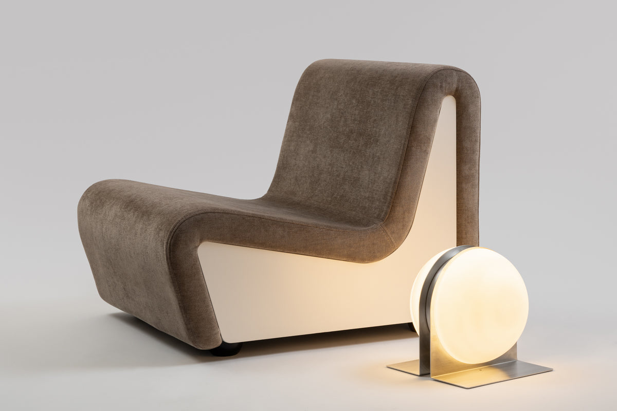 Dialogue lounge chair in Clay grey fabric with white lacquered wood side panels, with a lit Jumelles table lamp in brushed aluminium placed beside it