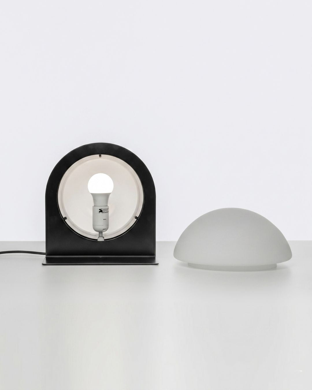Front view of the black “Jumelles” table lamp, disassembled, with its bulb exposed and its glass globe placed next to it